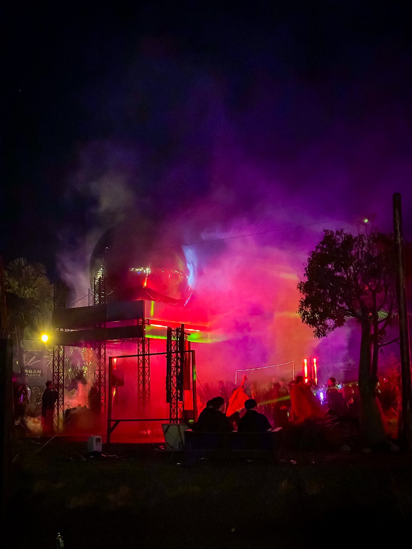 A crowd dancing in red and purple smoke beneath an outdoor stage at night